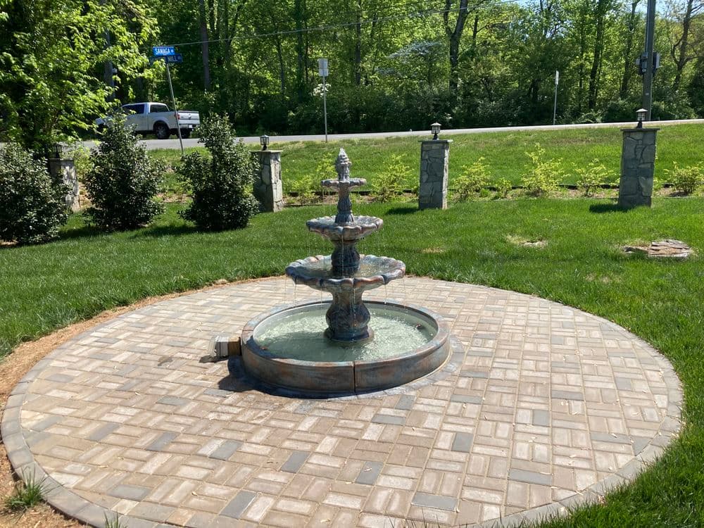 Three-tiered decorative fountain surrounded by landscaped grass and stone pathway.
