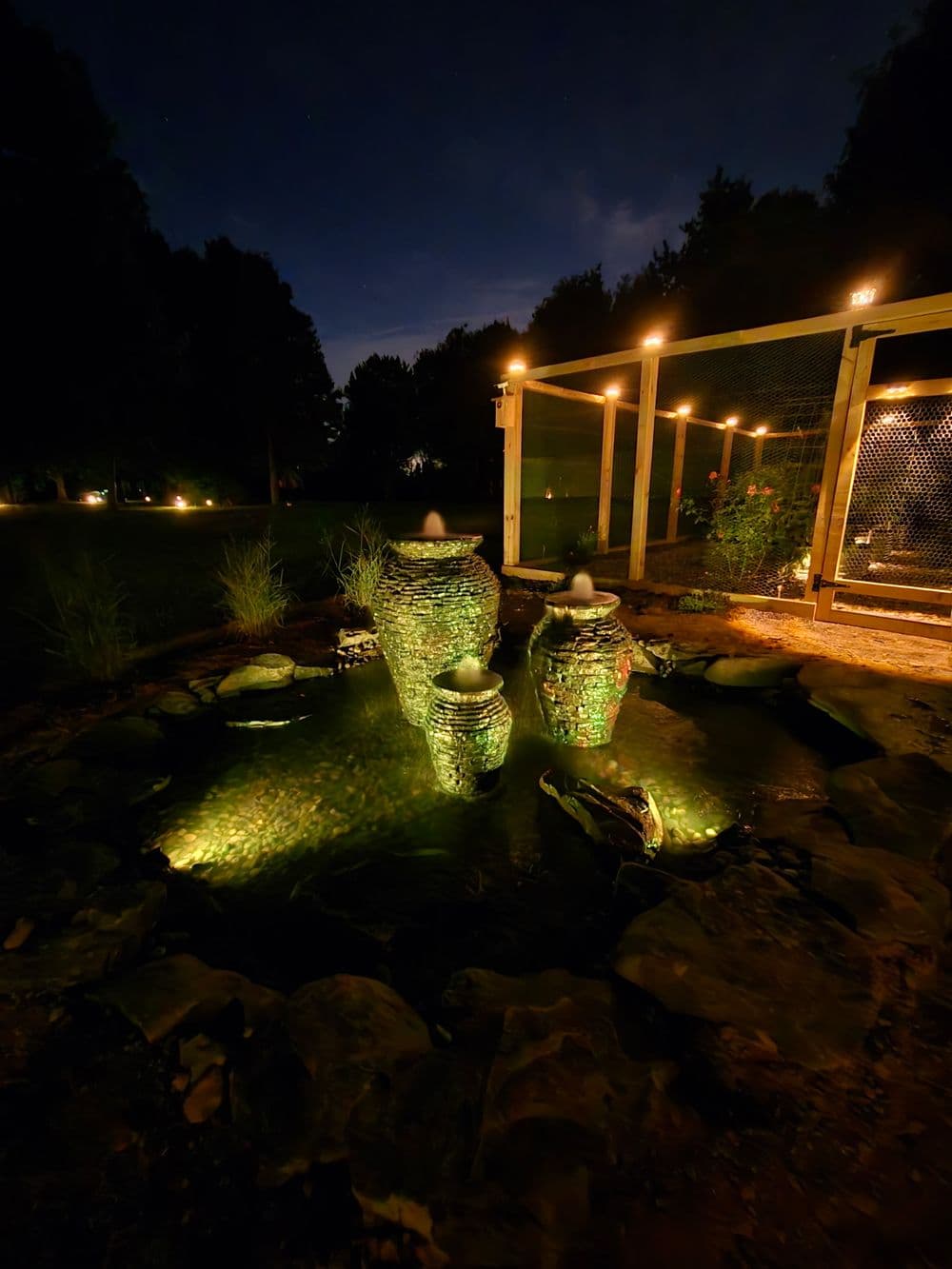 Decorative nighttime water fountain with green lights and surrounding landscaping.