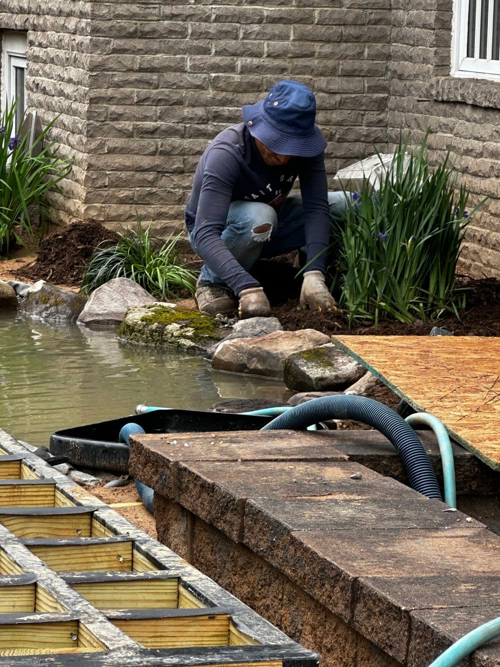 Person gardening by a pond, planting flowers and adjusting landscape features.