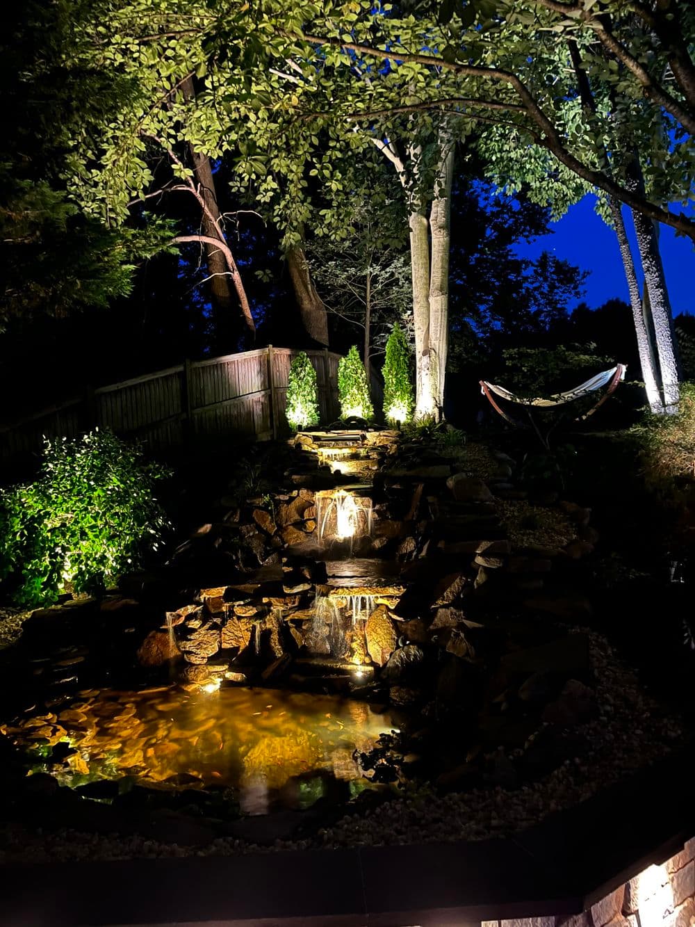 Serene backyard waterfall feature illuminated at night with surrounding greenery.