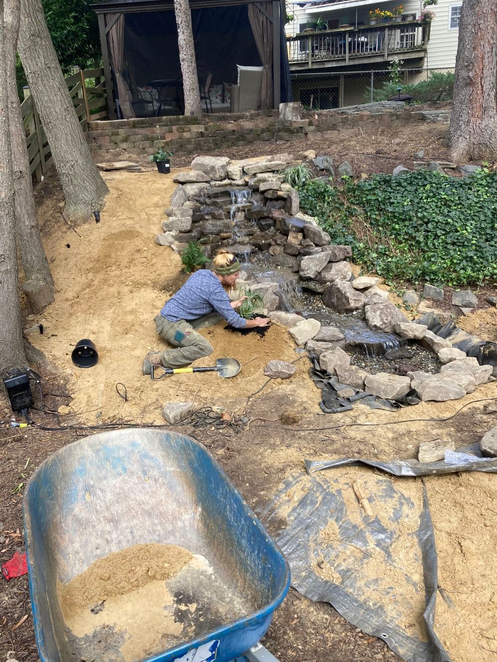 Person landscaping a garden with rocks and plants by a small water feature in a backyard.