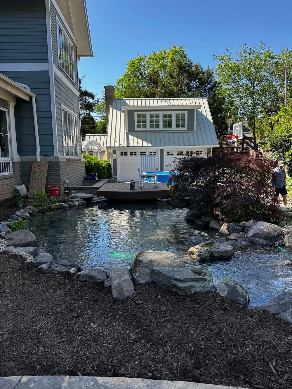 Lush backyard with a pond, rocks, and a deck, featuring a modern house in the background.