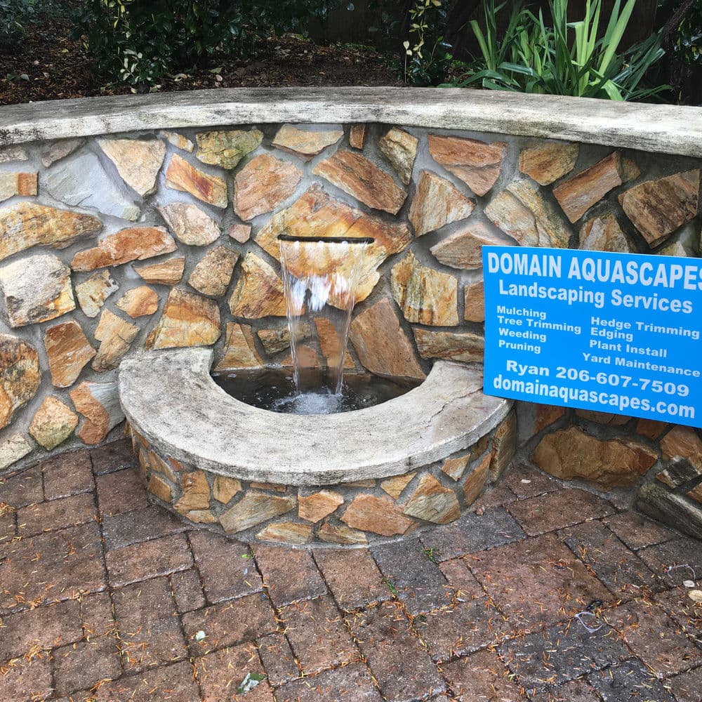 Decorative stone fountain with a blue sign for Domain Aquascapes landscaping services.